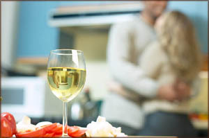 Valentines Day idea for homecooking. Photo of a glass of white wine in kitchen and a couple hugging in the background.